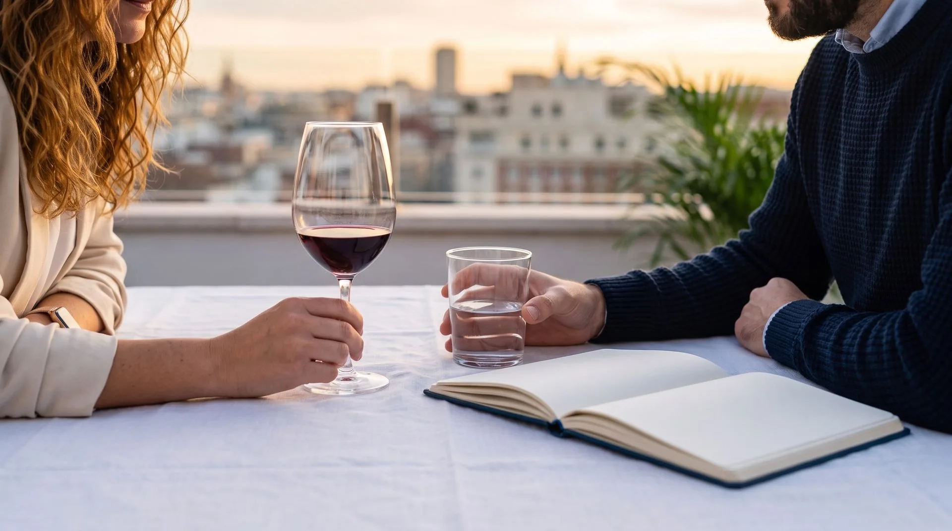 Sobremesa en una terraza al atardecer: manos, copas y un cuaderno abierto sobre el mantel